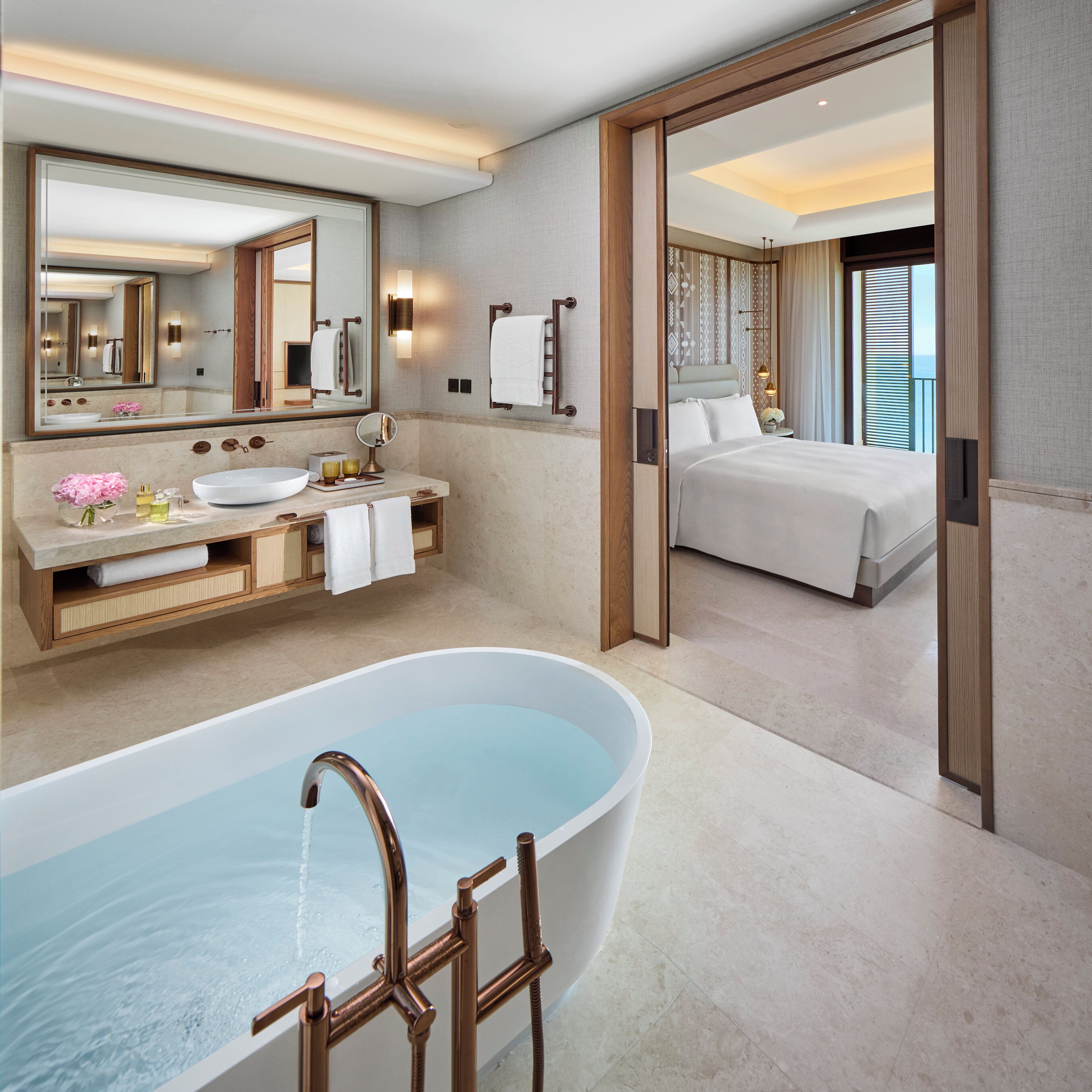 A modern bathroom with a freestanding bathtub, a large mirror, a vanity with a sink, and a bedside table with a lamp.