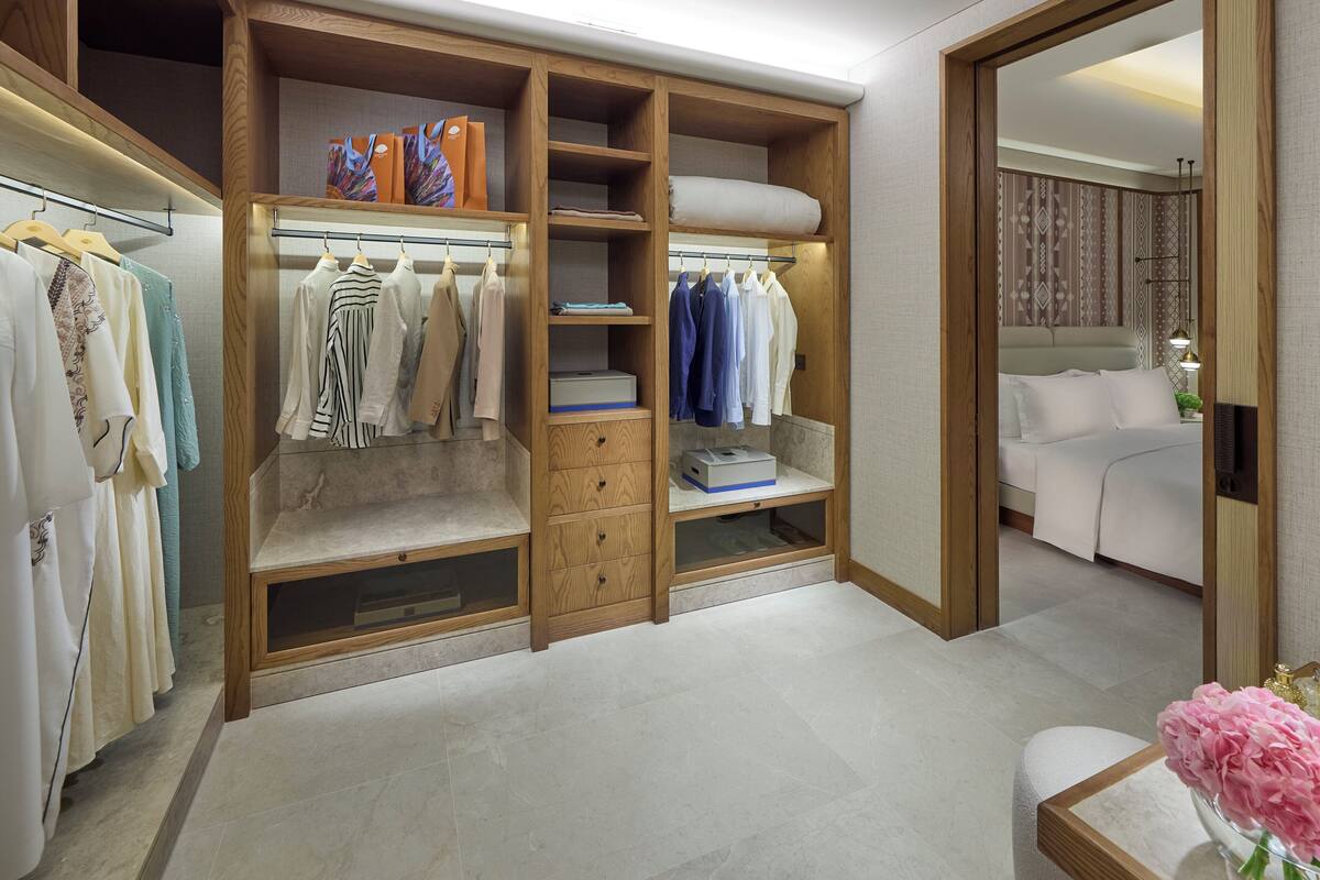 A walk-in closet with multiple shelves and drawers, a bed, and a bathroom with a sink and mirror.