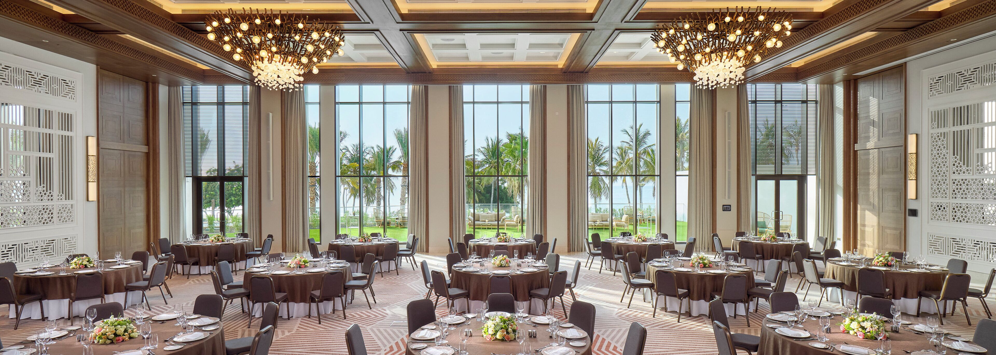 A spacious banquet hall with round tables, elegant chandeliers, and large windows offering a view of the outdoors.