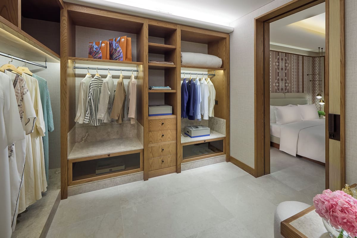 A walk-in closet with multiple shelves and drawers, a bed, and a bathroom with a sink and mirror.