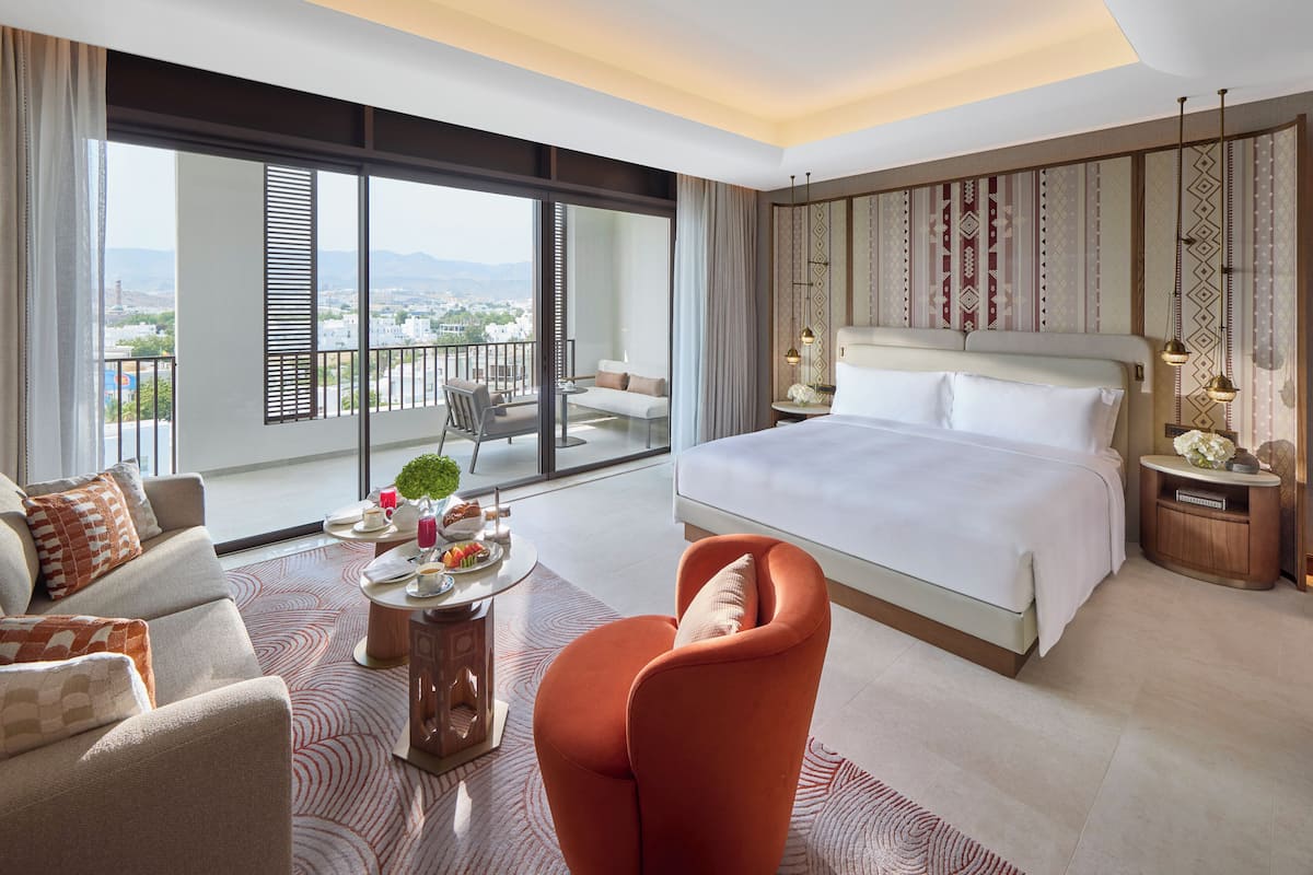 A modern hotel room with a large bed, a balcony with a view, a sitting area, and a dining table.