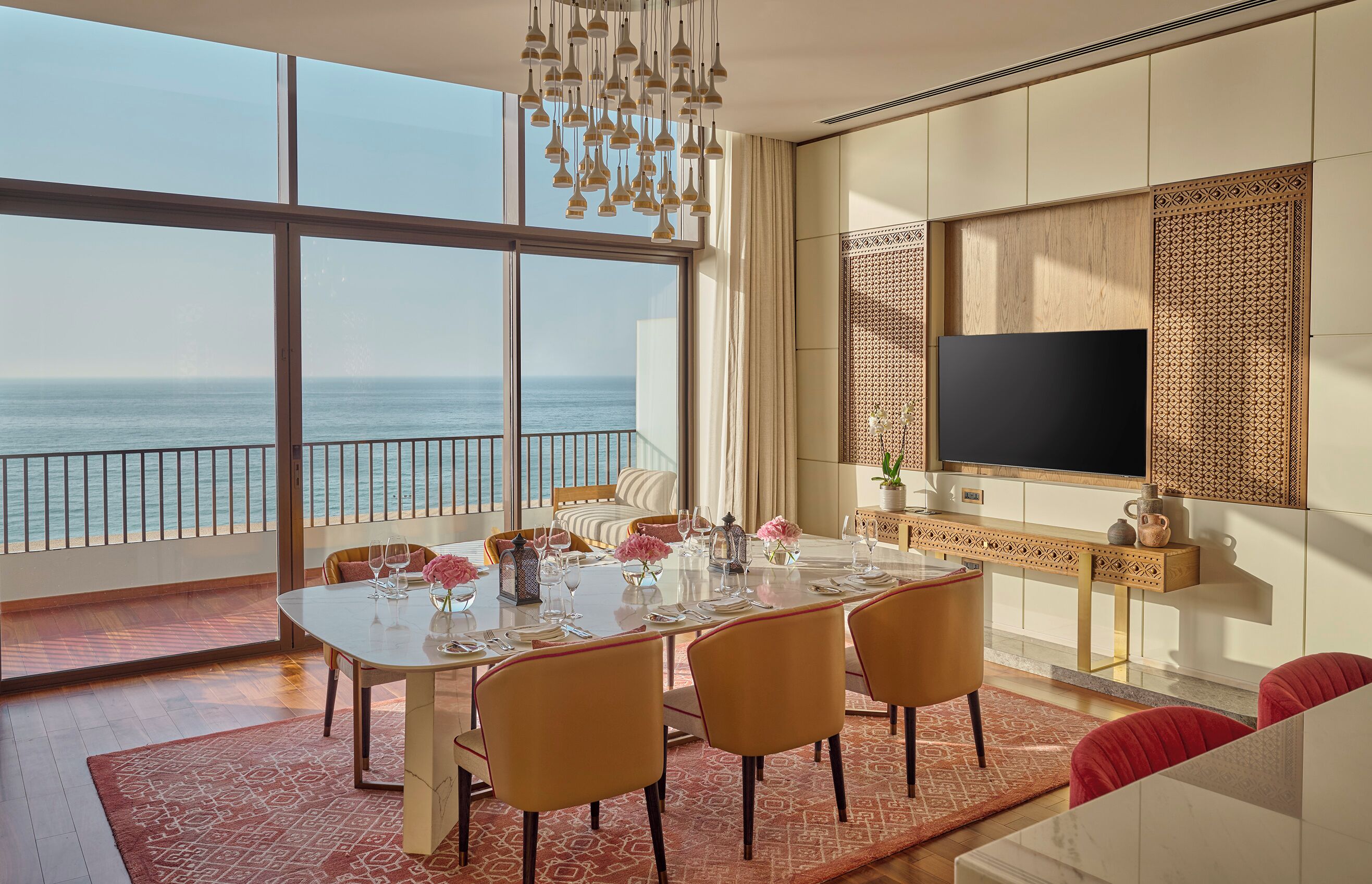 A dining area with a large table set for six, a television mounted on the wall, and a view of the ocean through large windows.