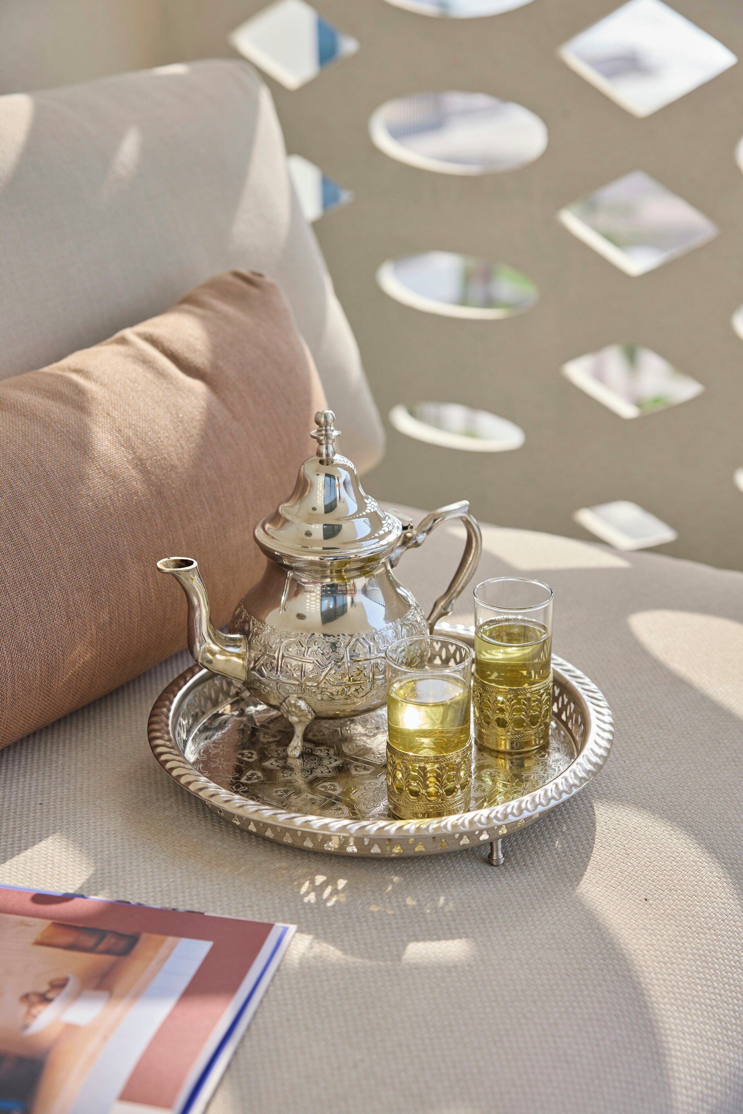 A tray with a teapot and two glasses of tea, placed on a table with a cushion and a magazine.