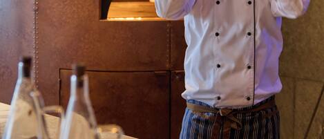 Un chef con uniforme blanco y pantalones a rayas, de pie frente a un horno de ladrillo.