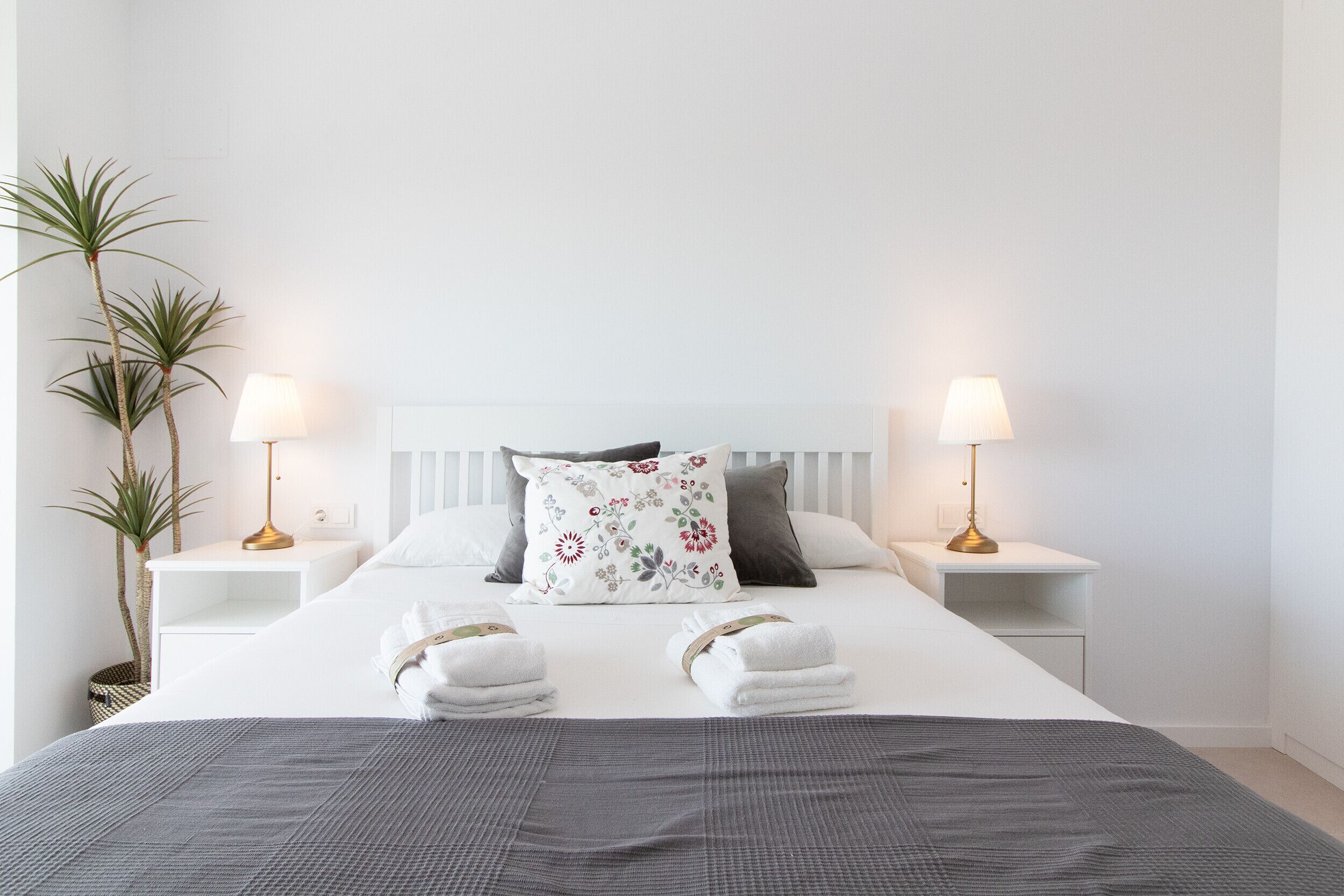 Un lit bien fait, avec du linge de lit blanc, deux lampes de chevet, un coussin à motifs floraux et un plaid gris.