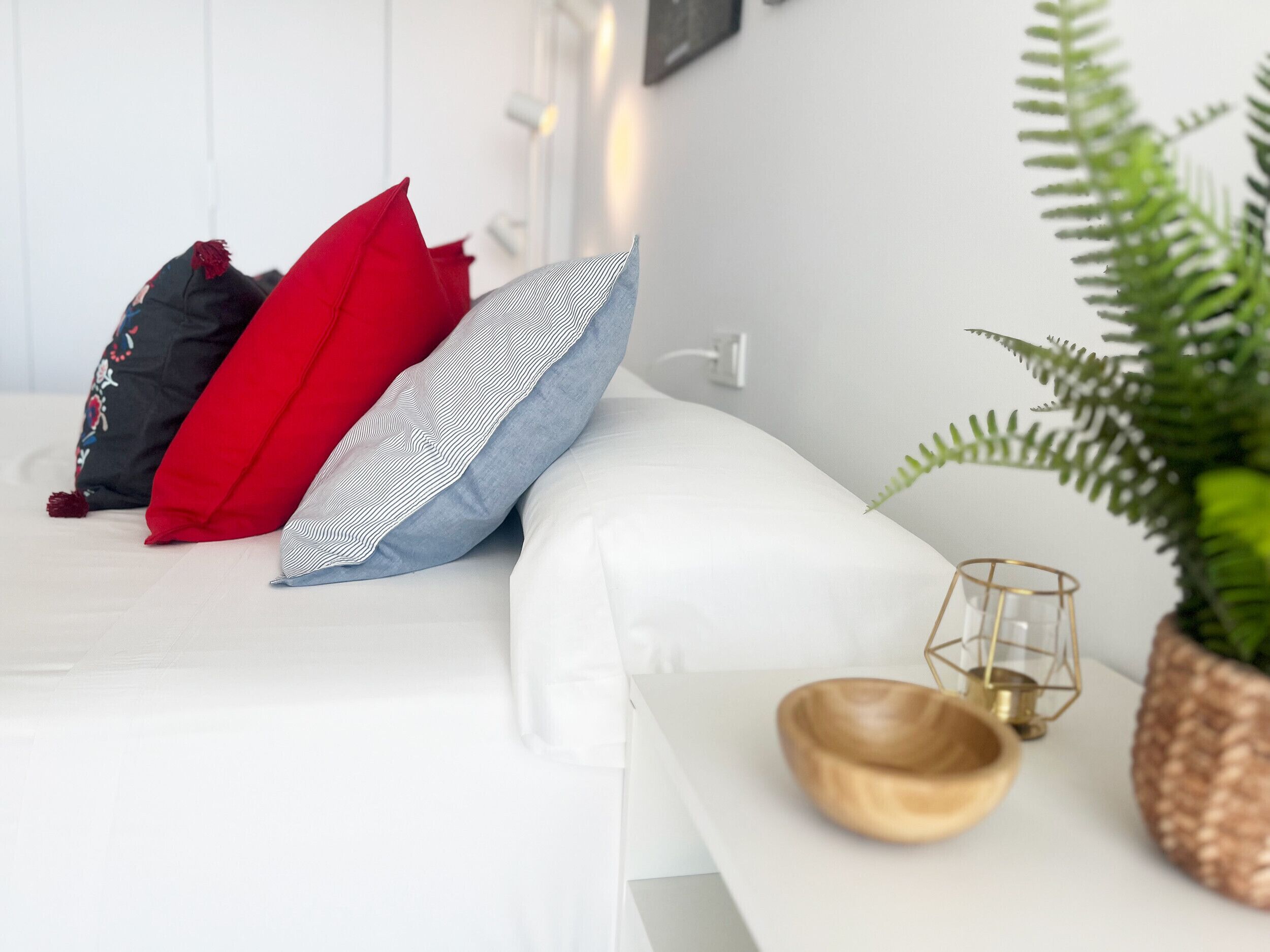 Un dormitorio ordenado con una cama, almohadas, una mesita de noche con una planta, una vela y un cuenco decorativo.
