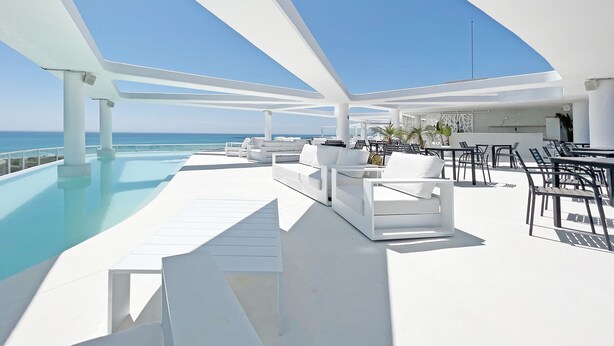Un área de piscina moderna al aire libre con sillas y mesas de lounge blancas, una vista despejada del océano y una terraza amplia.