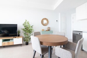 A modern dining area with a round table, four chairs, a television, a potted plant, a decorative mirror, and a kitchen area with a dishwasher.