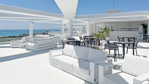 Un espacio lounge moderno al aire libre con mobiliario blanco, incluyendo sofás y mesas, con vista a un cielo azul despejado y una costa distante.