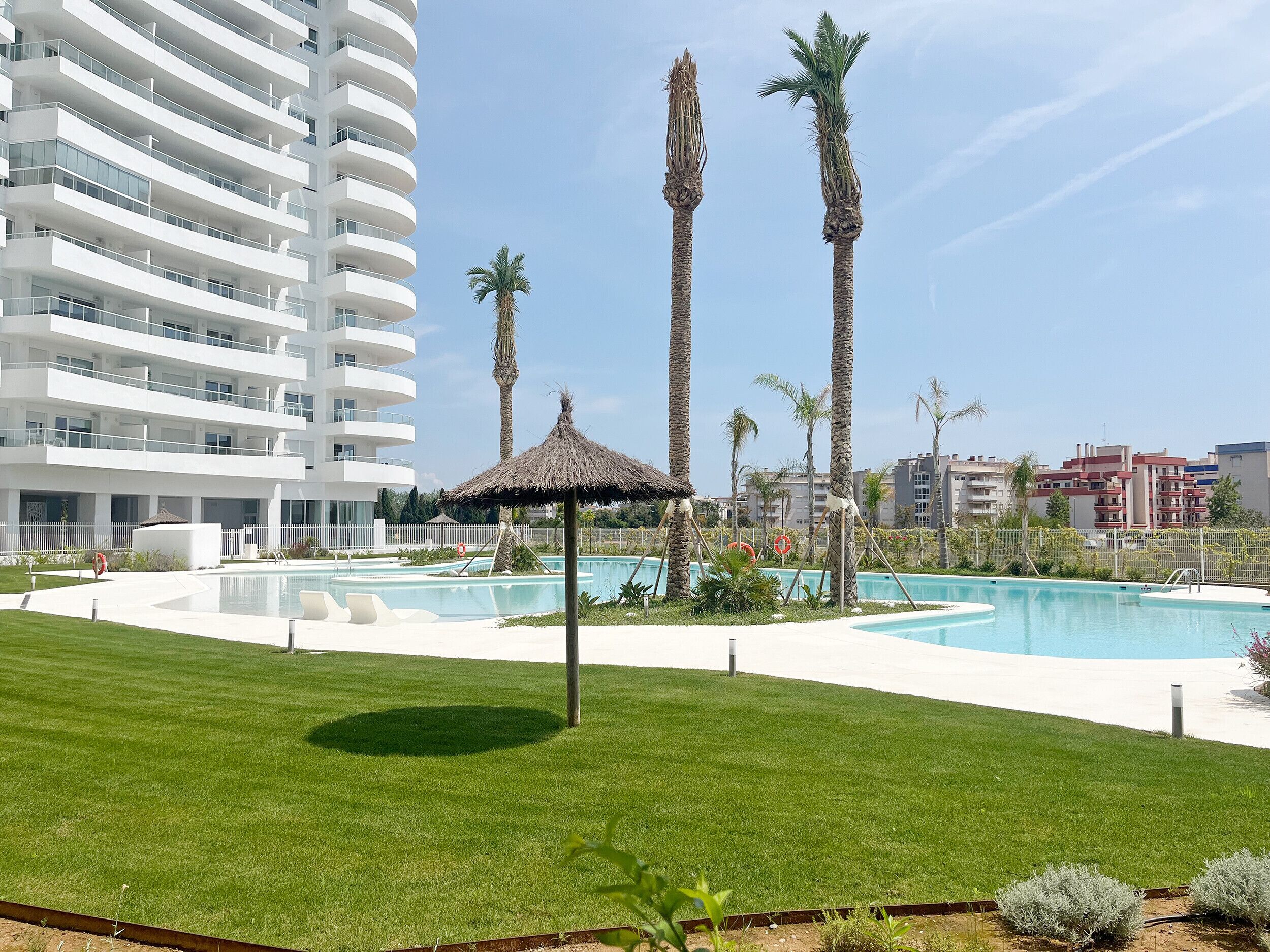 A modern hotel with a pool area, palm trees, and a thatched-roof gazebo.