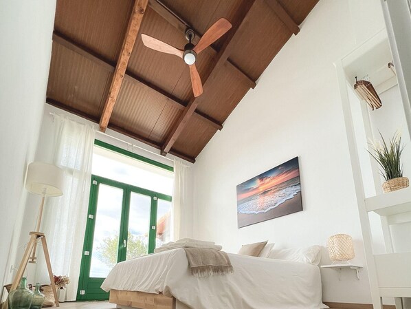 A bedroom with a bed, a ceiling fan, a painting, and a view of the outdoors.