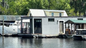 Una casa flotante con balcón, un pequeño porche y una zona de embarcadero con barcos.