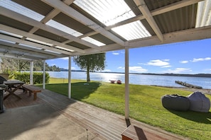 A covered outdoor area with a wooden deck, picnic table, and bean bag chairs, overlooking a lake with a dock.