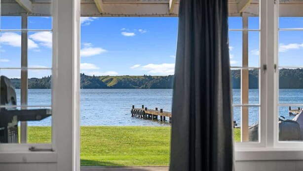 A room with large windows offering a view of a lake, a wooden dock, and a grassy area.