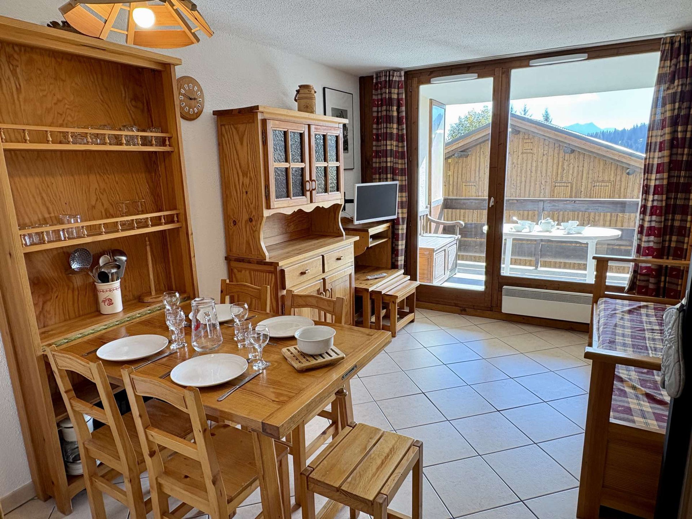 A wooden dining area with a table set for four, a television, and a view of a wooden cabin through a glass door.