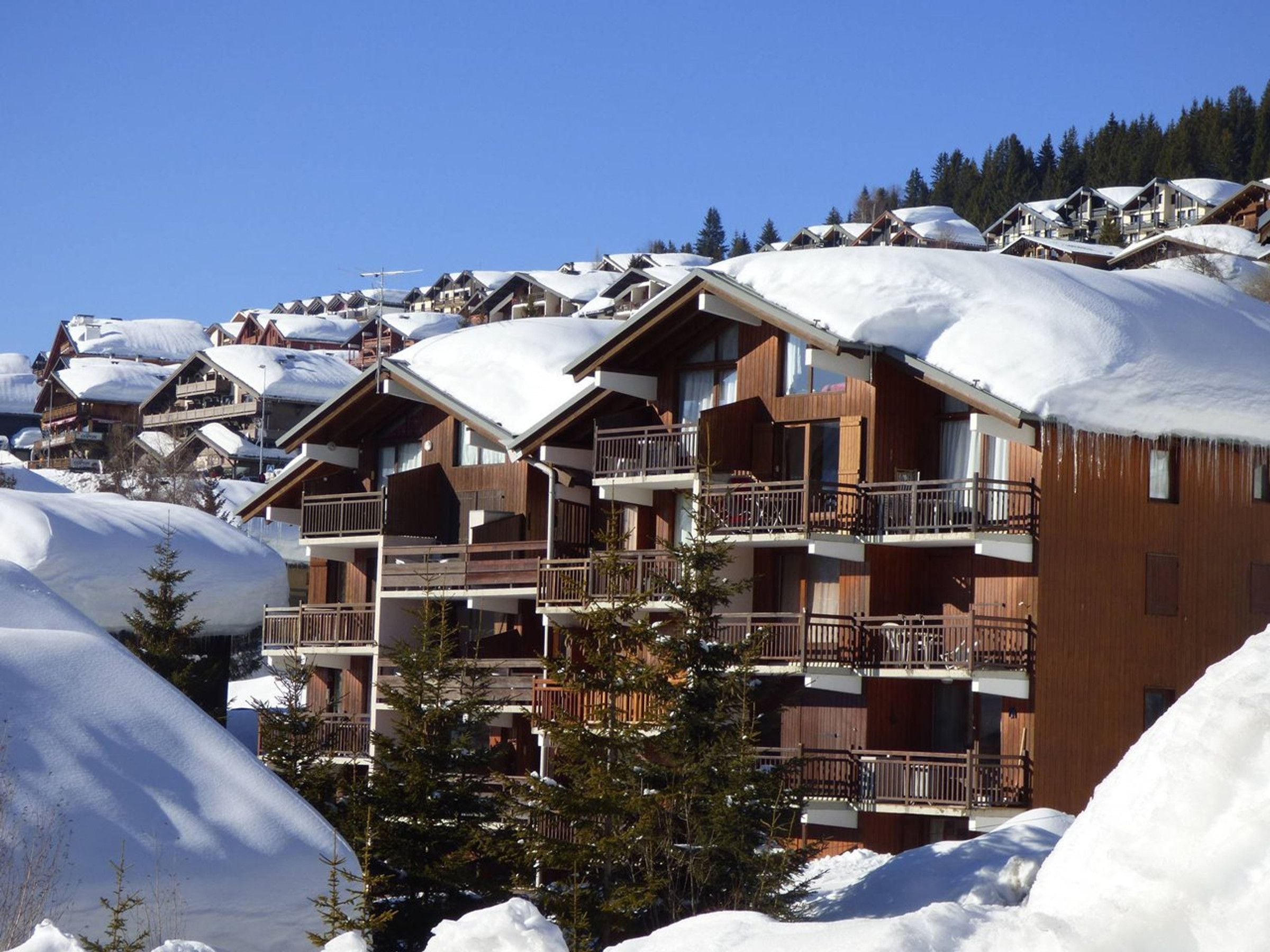 Ein mehrstöckiges Holzhaus mit Balkonen, umgeben von schneebedeckten Bäumen und anderen Gebäuden.