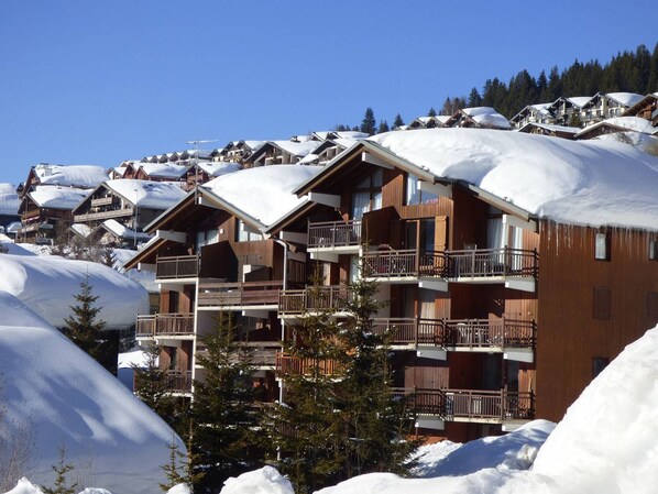 A multi-story wooden building with balconies, surrounded by snow-covered trees and other buildings.