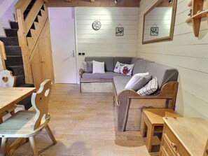 A cozy room with a wooden staircase, a grey sofa with cushions, a wooden dining table with chairs, and a small wooden cabinet.