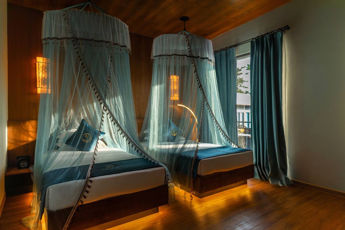 A room with two beds, each with a canopy, wooden flooring, and a view of the outdoors.