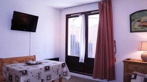 Une pièce comprenant une table, une chaise, un téléviseur fixé au mur, une fenêtre avec des rideaux et un tableau accroché au mur.