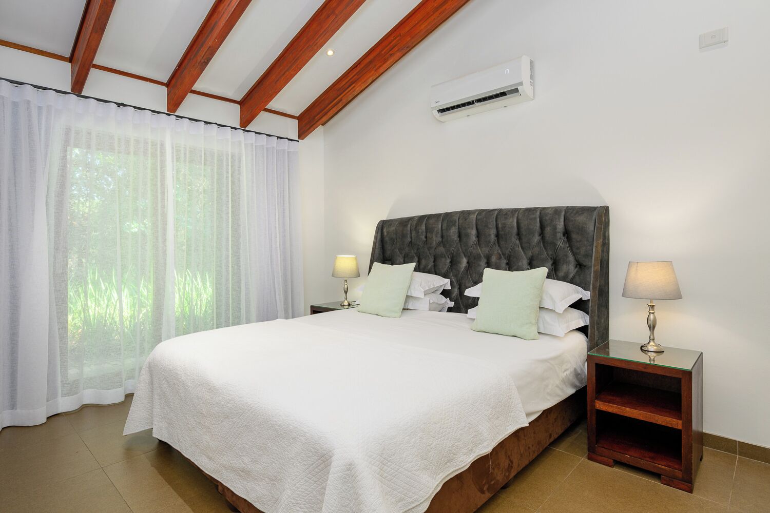 A bedroom with a bed, two bedside tables with lamps, a window with sheer curtains, and an air conditioning unit.