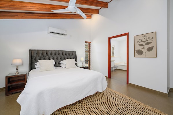 Une chambre avec un grand lit, un ventilateur de plafond, un appareil de climatisation, une table de chevet, une lampe de chevet, un tapis tissé et une salle de bain visible à travers une porte.
