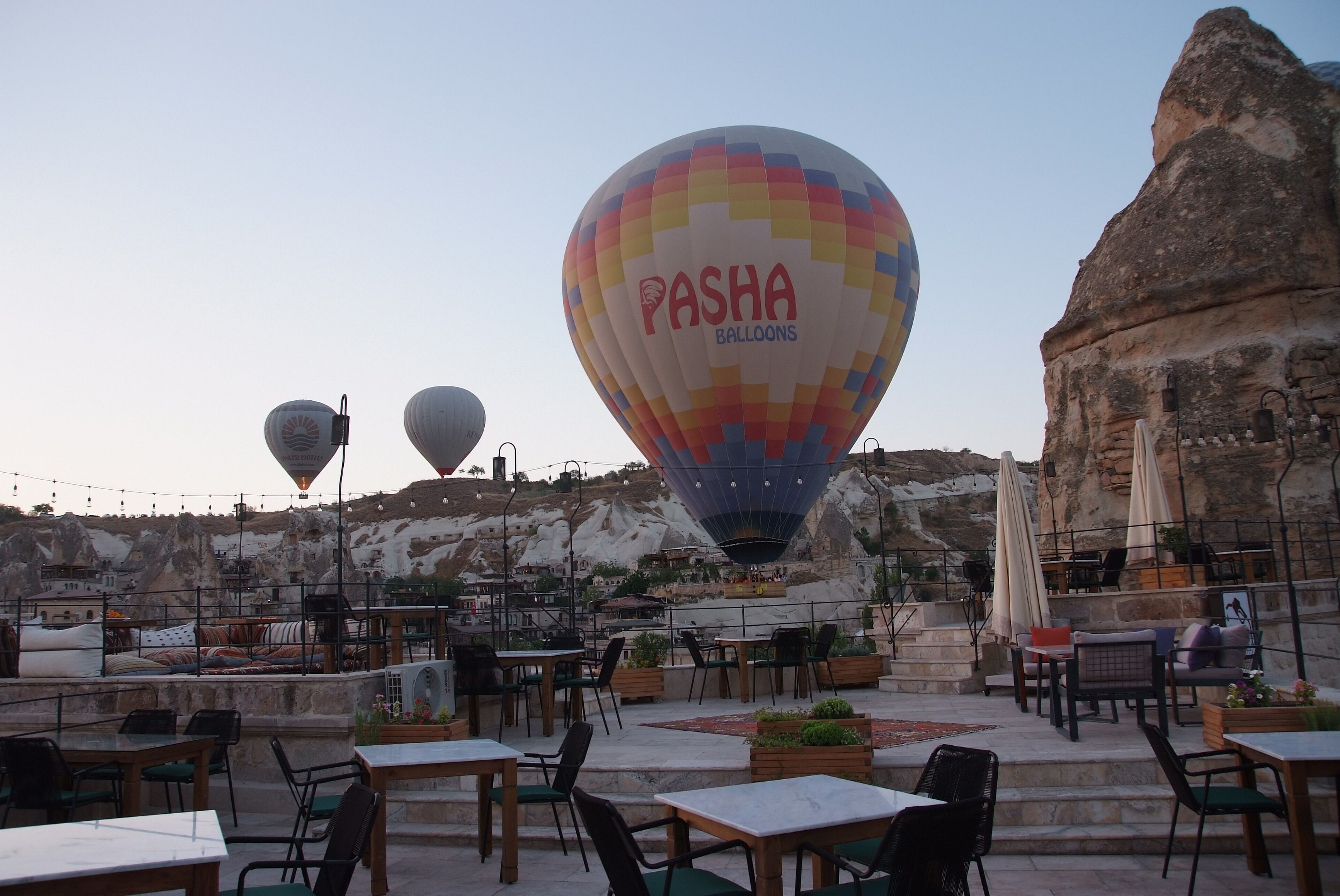 Een terras op het dak met tafels en stoelen, een luchtballon met de branding 'Pasha Balloons' en een rotsachtig landschap op de achtergrond.