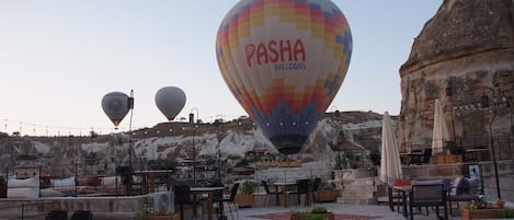 Een terras op het dak met tafels en stoelen, een luchtballon met de branding 'Pasha Balloons' en een rotsachtig landschap op de achtergrond.
