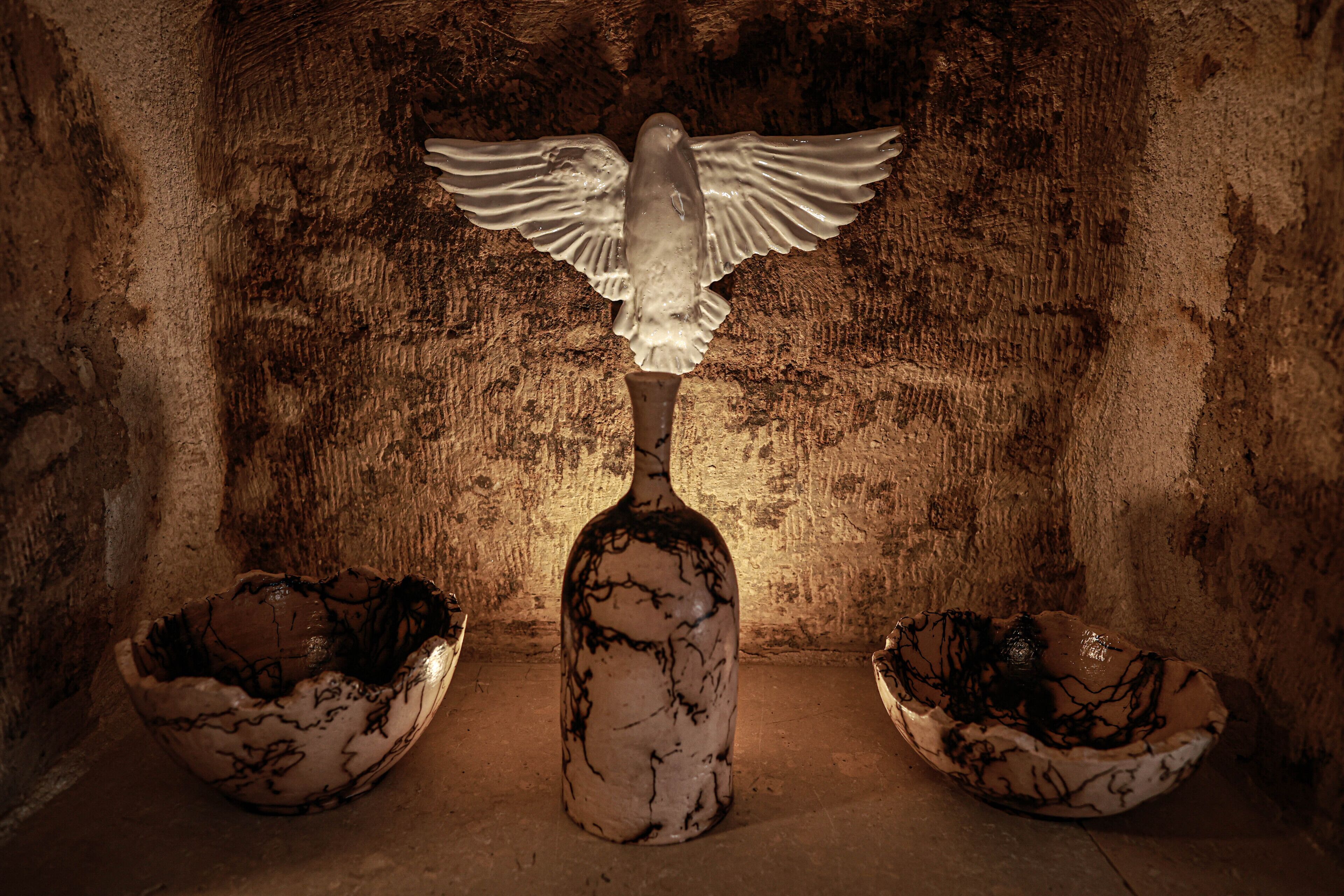 A sculpture of a bird with outstretched wings above a bottle, flanked by two bowls on a textured surface.