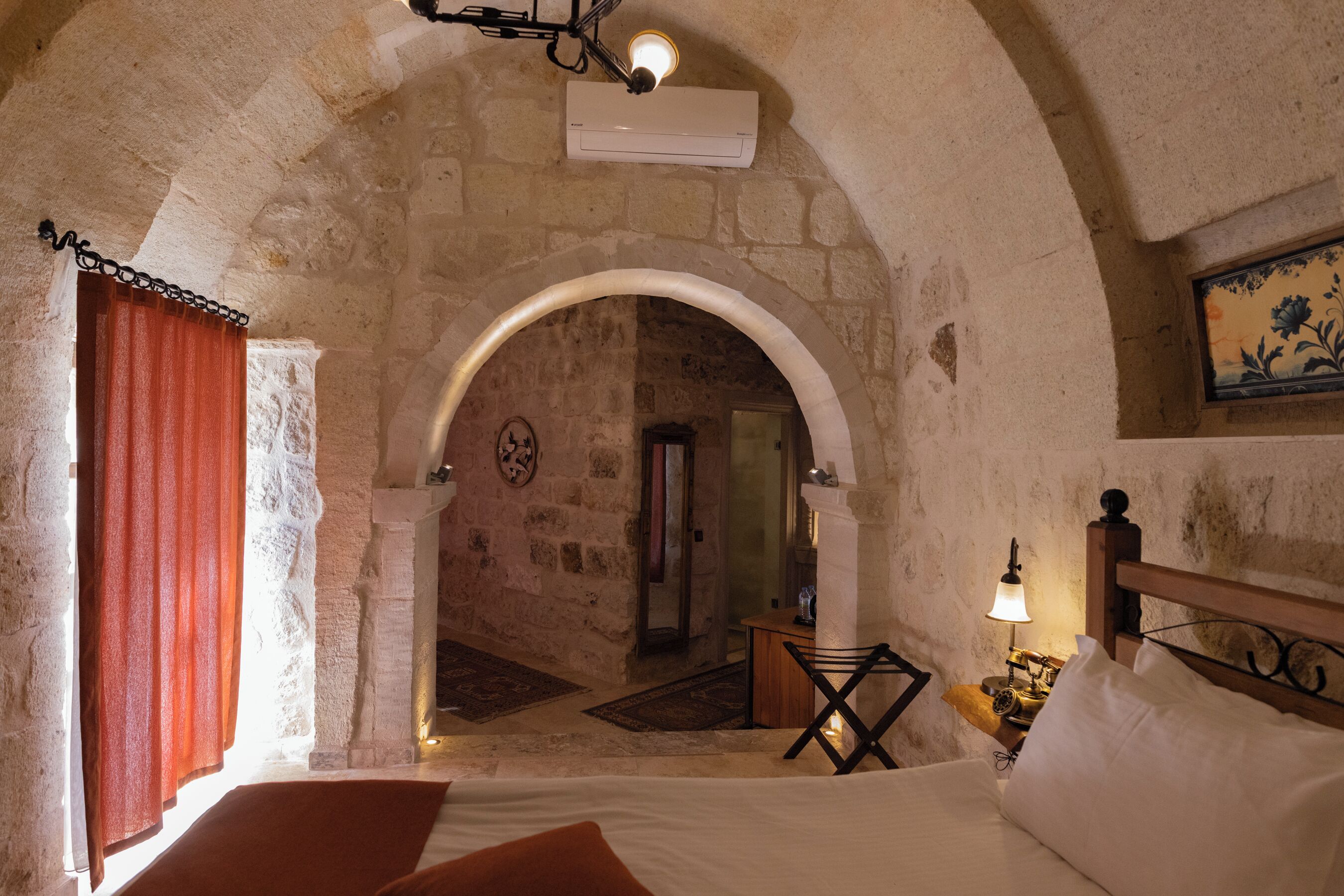 Un dormitorio con un arco de piedra, una cama con ropa blanca, una mesita de noche con lámpara y una cortina roja.