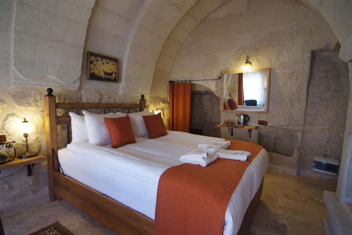 A bedroom with a wooden bed, white linens, a red throw blanket, a nightstand with a lamp, and a stone wall.