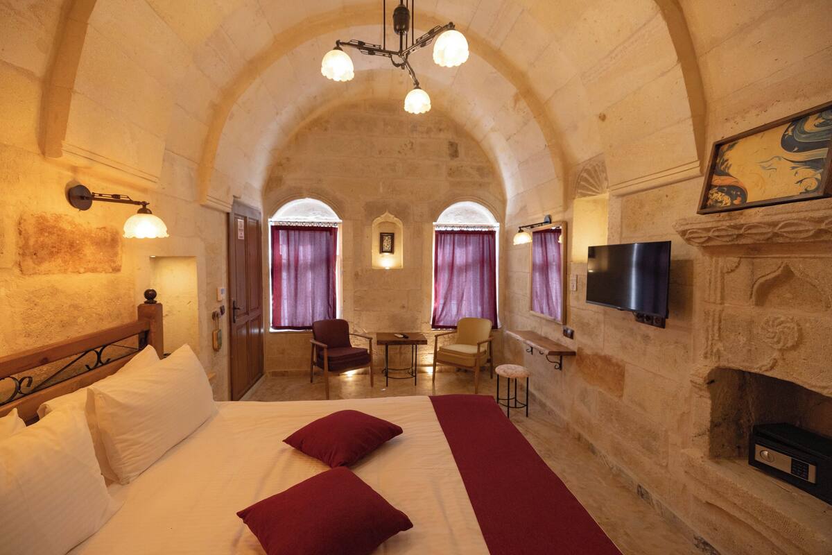 A room with a bed, two red pillows, a TV, a fireplace, and a stone wall.
