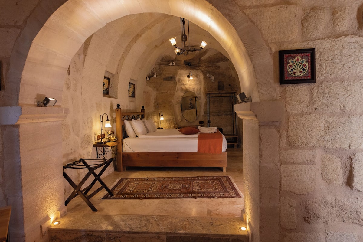 A cozy bedroom with a wooden bed, a small table, and a chair, under a stone archway.