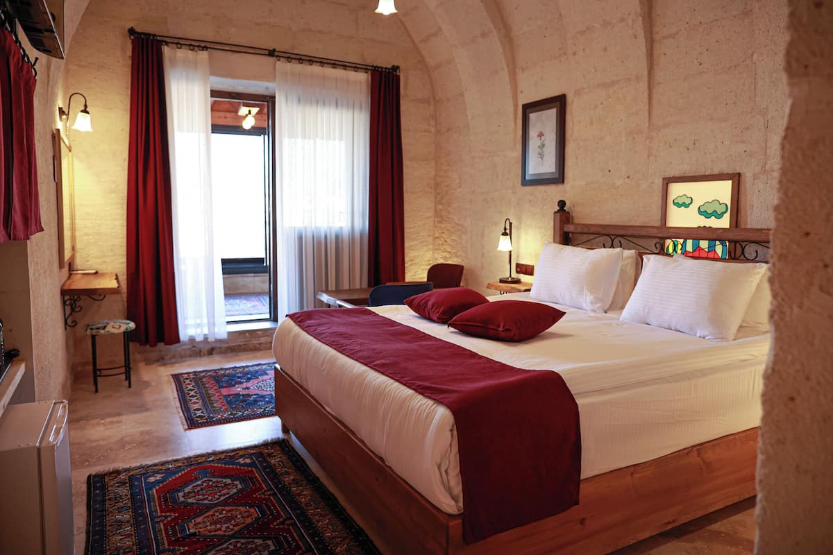 A bedroom with a wooden bed, red and white bedding, a patterned rug, a desk with a chair, and a view of the outside through a window with curtains.