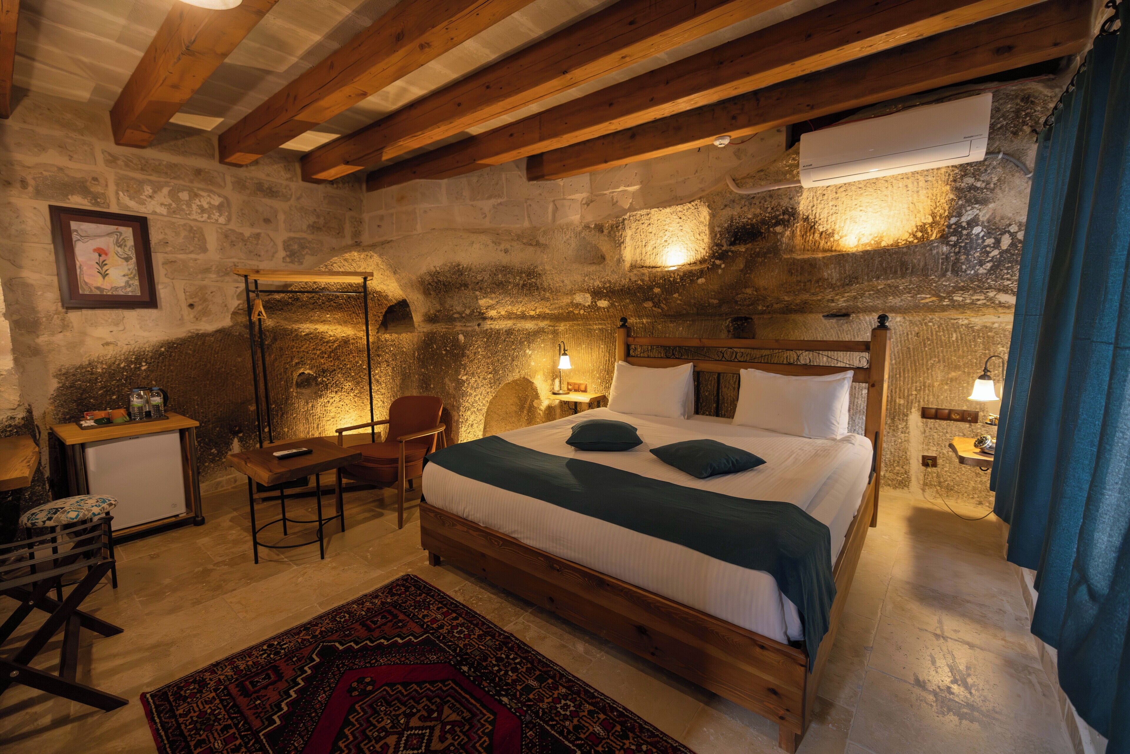 Ein rustikales Schlafzimmer mit Steinwand, Holzbalken, einem Bett mit Kissen, einem Schreibtisch mit Stuhl und einem kleinen Tisch mit Lampe.