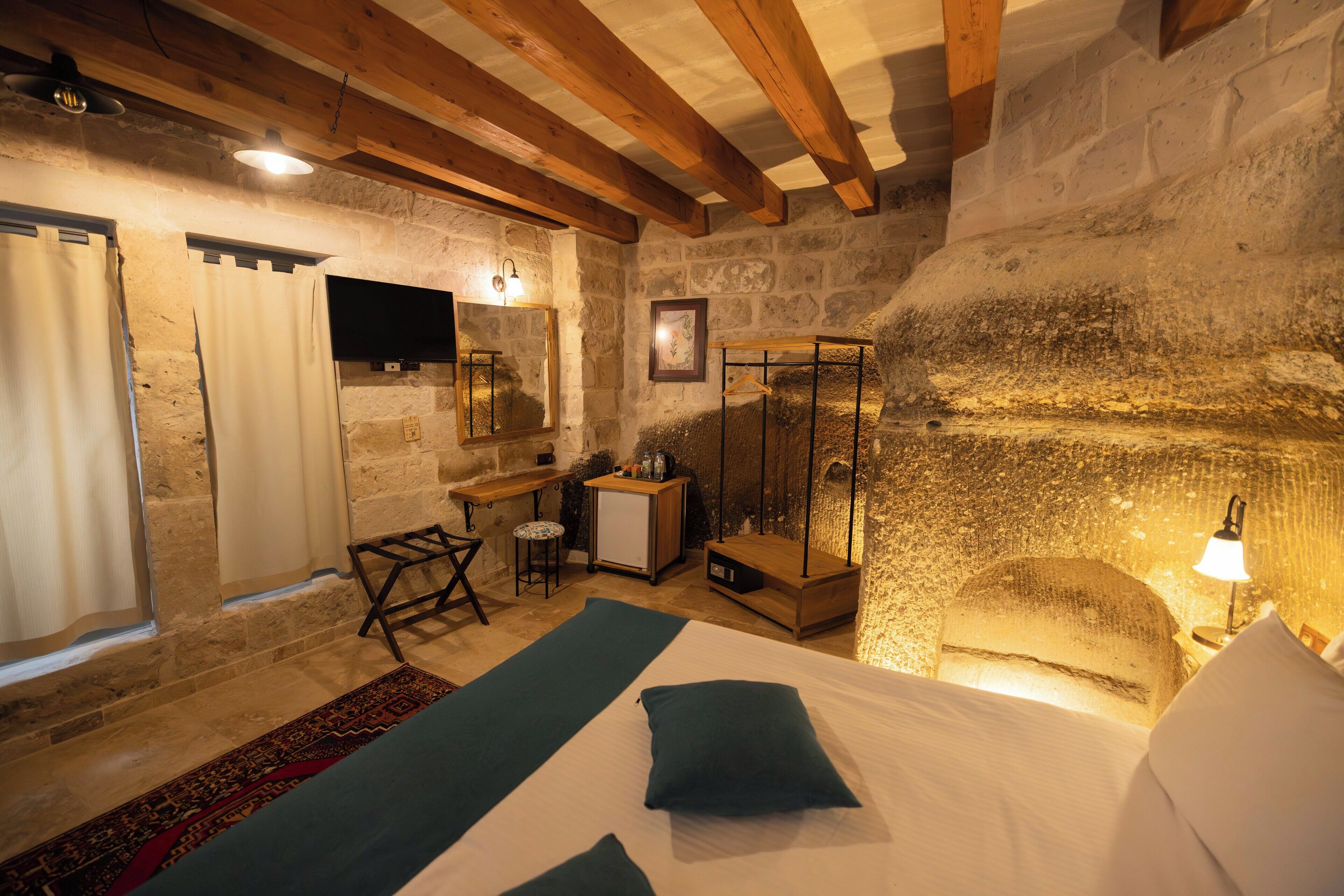 A rustic bedroom with a stone wall, wooden beams, a bed with pillows, a desk with a chair, a small table, and a mirror.