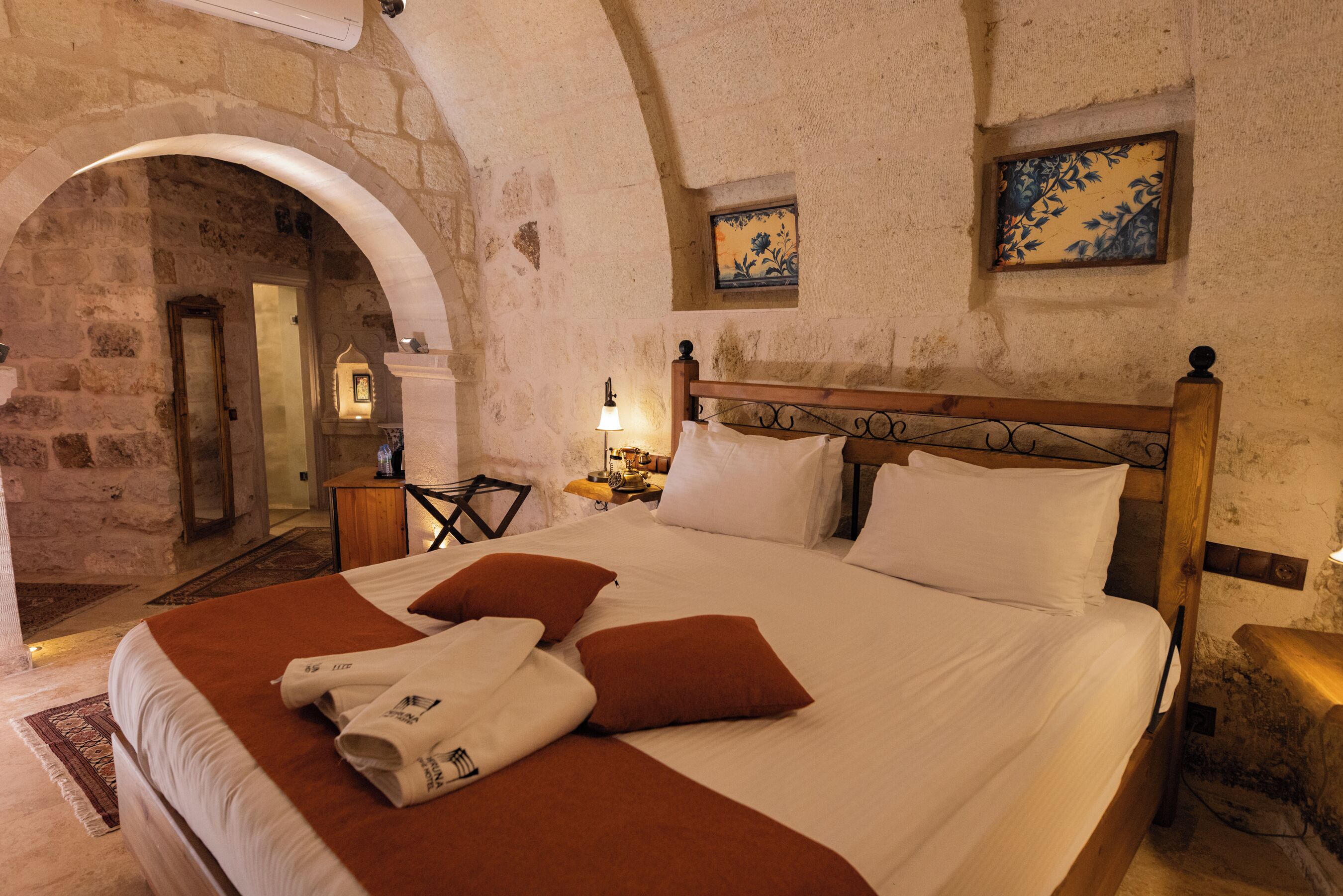 A bedroom with a stone wall, a wooden bed with white and brown bedding, a nightstand, a lamp, and a small table with a chair.