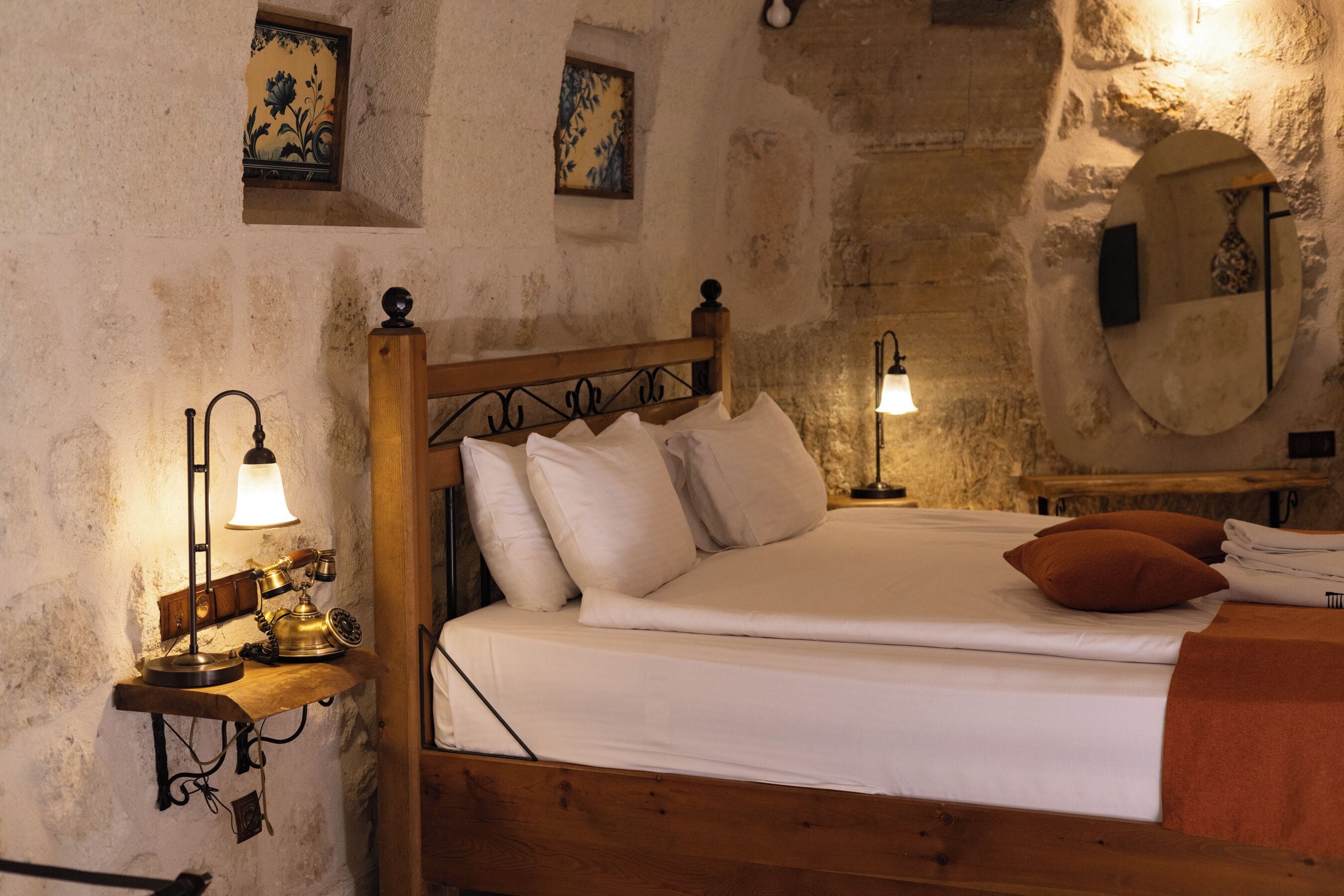 A wooden bed with white linens and a single brown pillow, a small bedside table with a lamp, and a wall-mounted mirror.