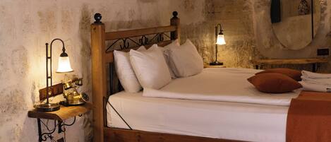 A wooden bed with white linens and a single brown pillow, a small bedside table with a lamp, and a wall-mounted mirror.