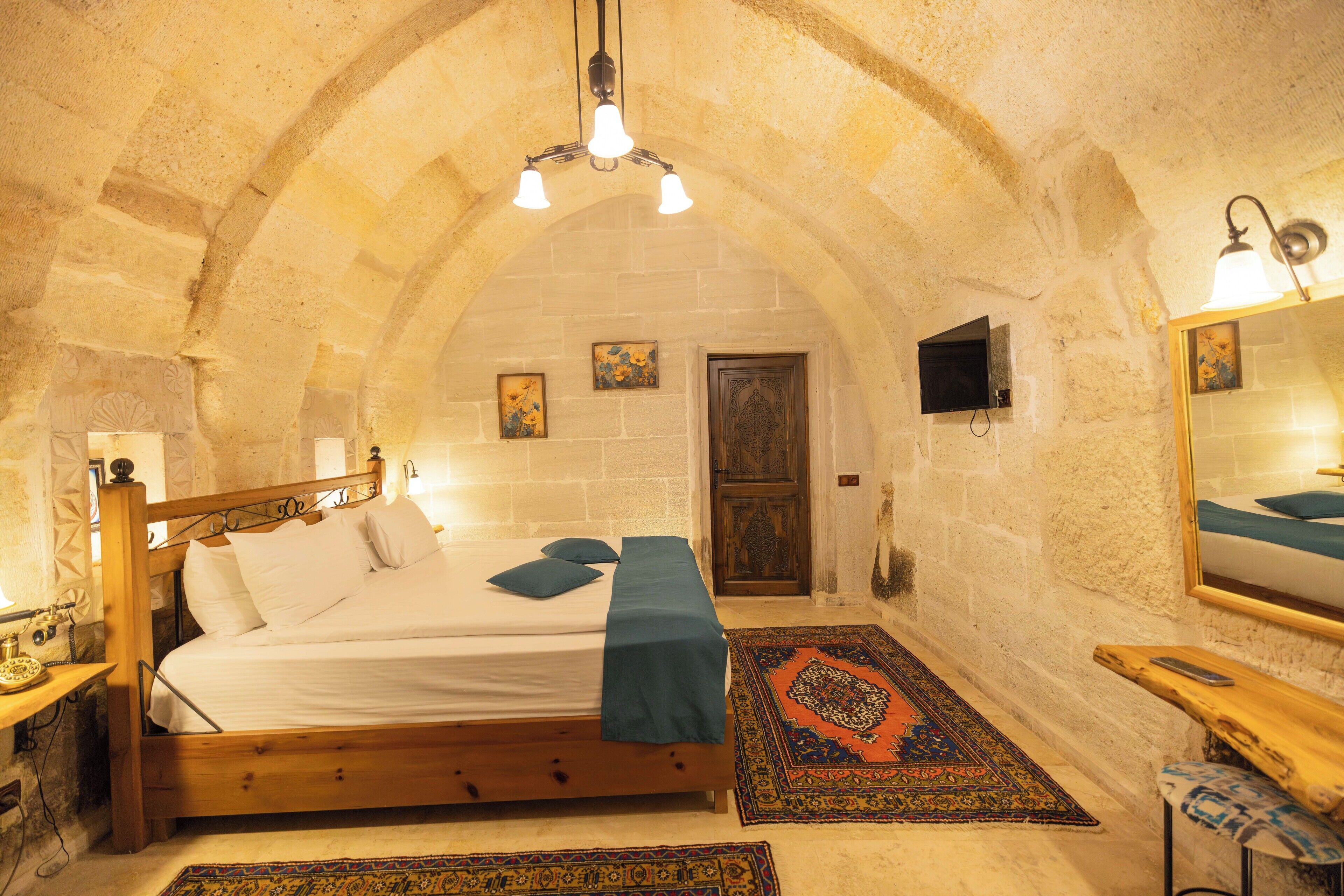 A bedroom with a wooden bed, a stone wall, a door, a mirror, and a small table with a lamp.