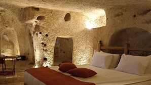 A room with a large bed, a wooden headboard, a nightstand, and a small table with a chair. The walls are made of stone with holes and a small window. - Peruna Cave (Nevsehir)