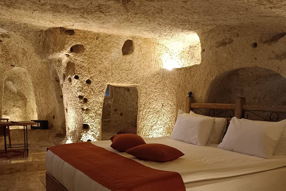 A room with a large bed, a wooden headboard, a nightstand, and a small table with a chair. The walls are made of stone with holes and a small window.