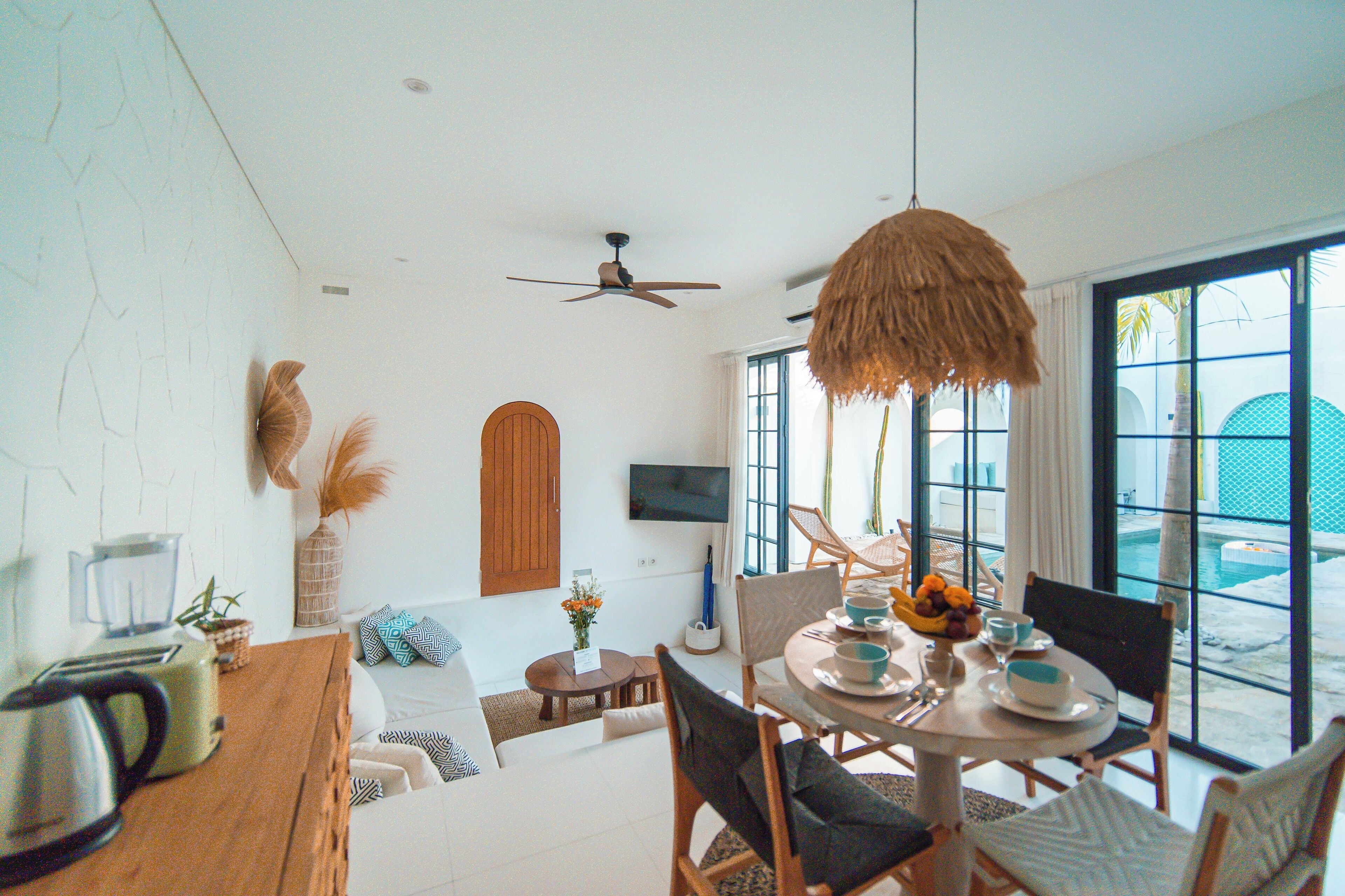 A modern living room with a dining area, a ceiling fan, a TV mounted on the wall, and large windows offering a view of the outdoors.