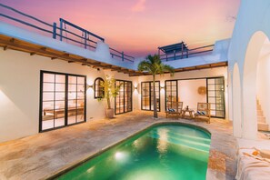 Zona de piscina con vista al atardecer, una palmera y un espacio de descanso con sillas y una mesa.