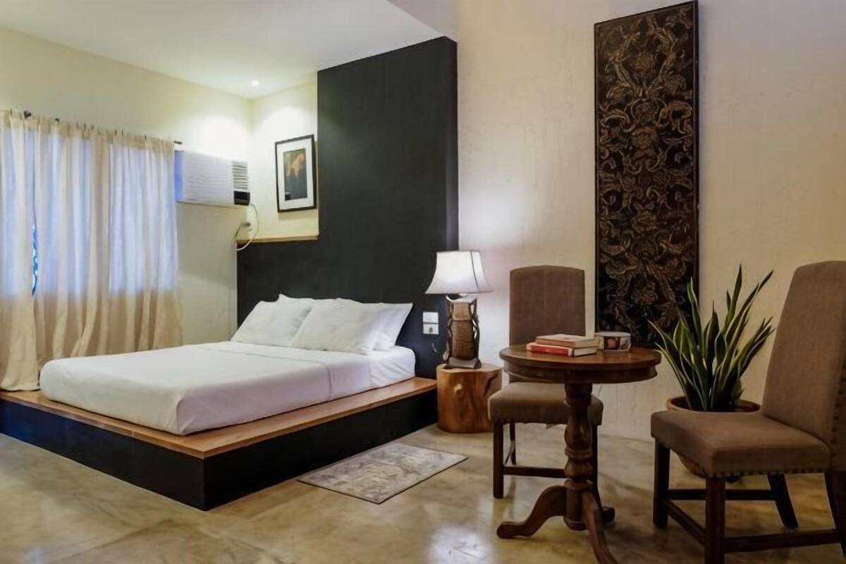 Habitación de hotel con cama, una silla, una mesita, una planta en maceta y un adorno en la pared.