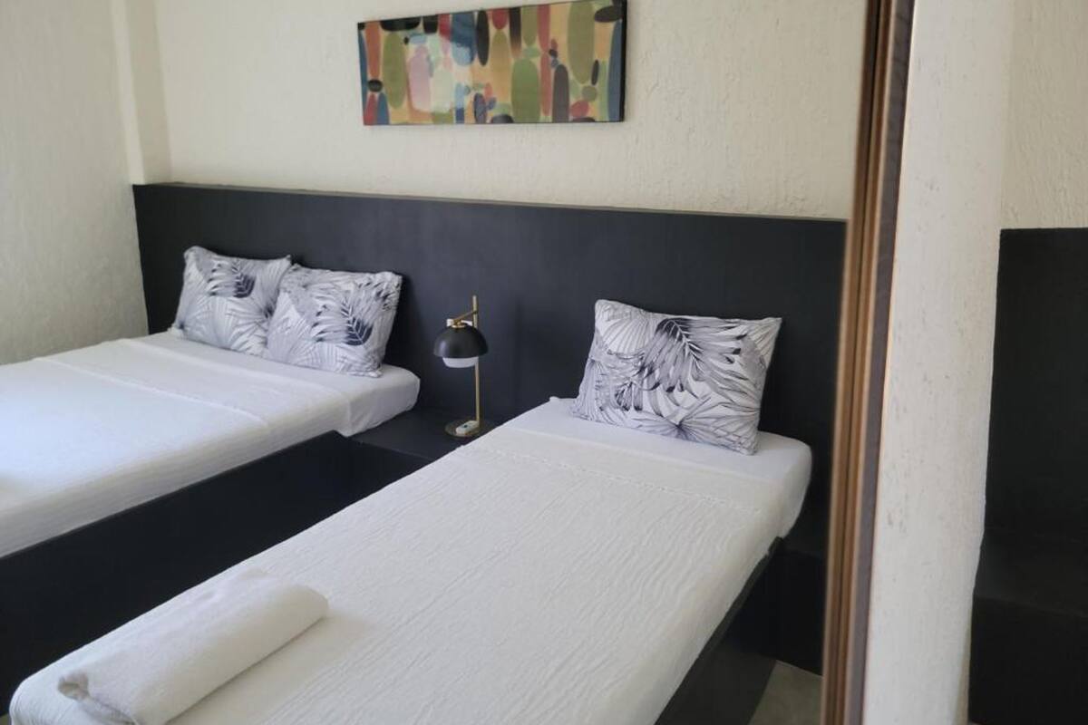 Una habitación de hotel con dos camas individuales, cada una con cubrecama blanco y almohada estampada con motivos vegetales. Un cabecero negro separa las camas, y un cuadro abstracto y colorido cuelga de la pared de arriba.