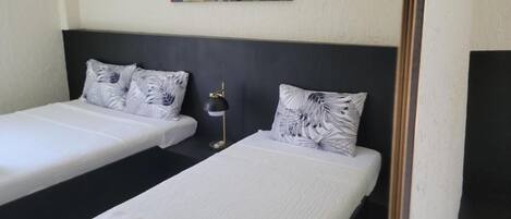 Una habitación de hotel con dos camas individuales, cada una con cubrecama blanco y almohada estampada con motivos vegetales. Un cabecero negro separa las camas, y un cuadro abstracto y colorido cuelga de la pared de arriba.