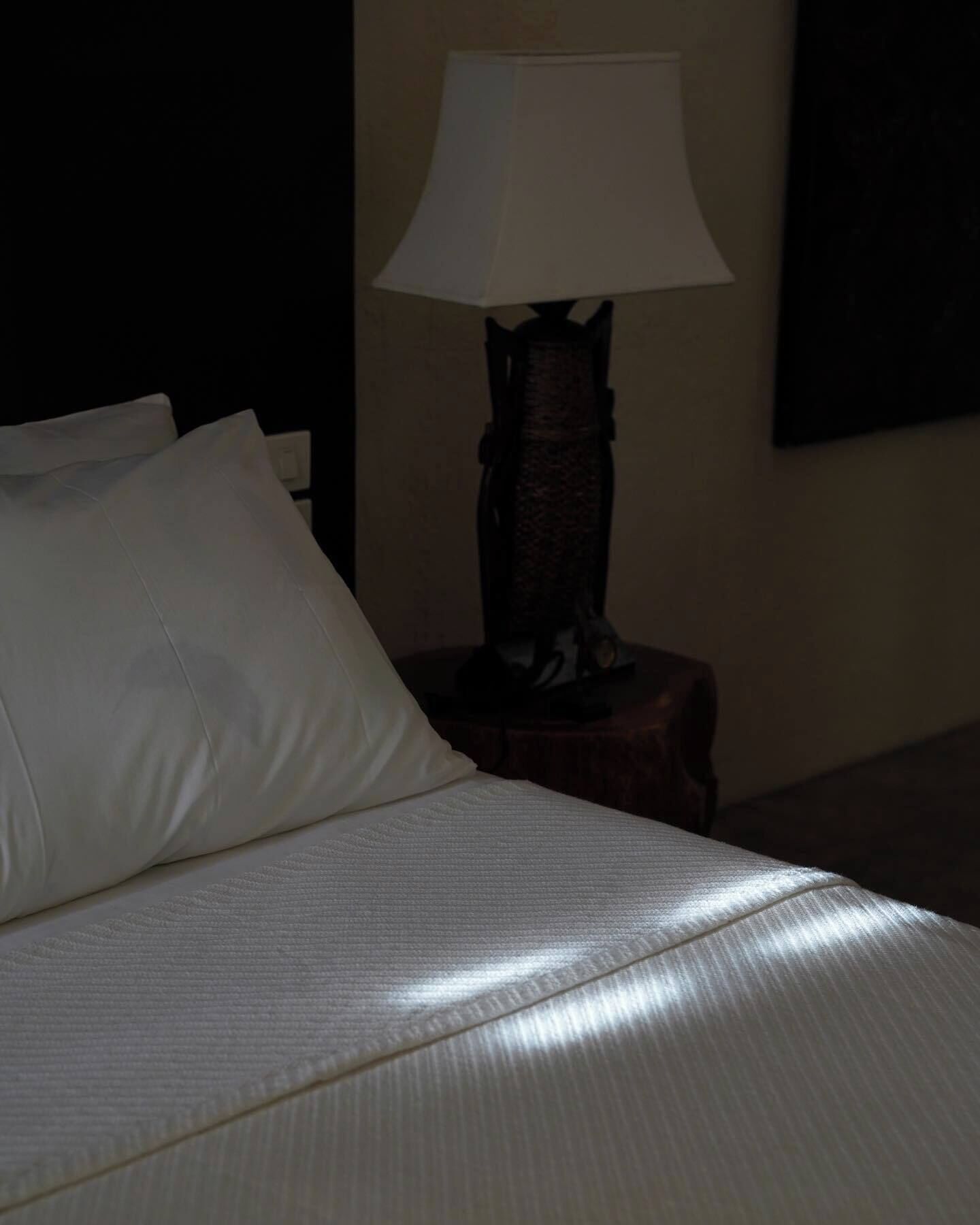 Una cama con un almohadón blanco y una manta texturizada, una mesita de noche de madera con una lámpara y una pared oscura.
