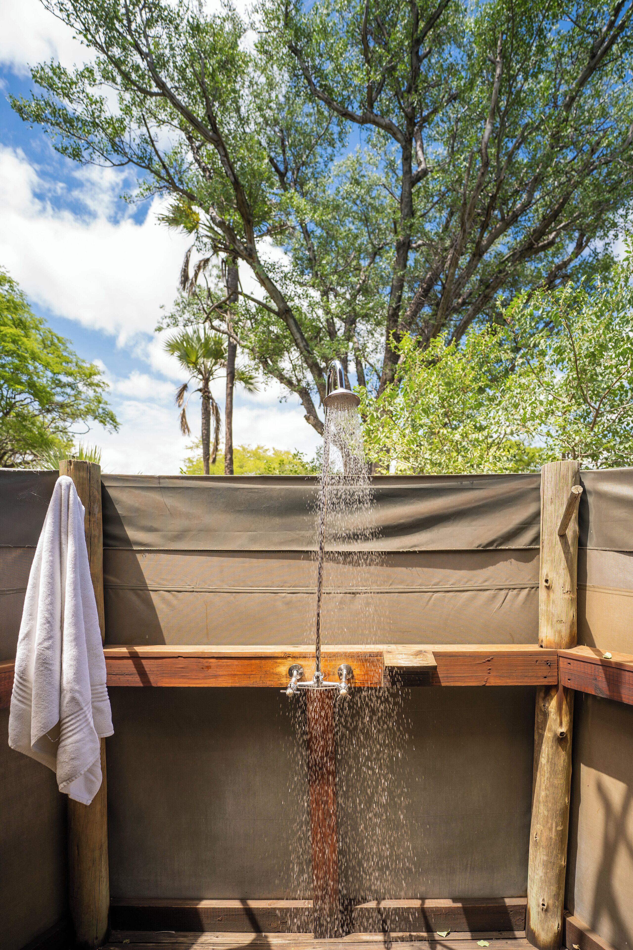 A wooden outdoor shower with a wooden bench, surrounded by a brown fence and trees.