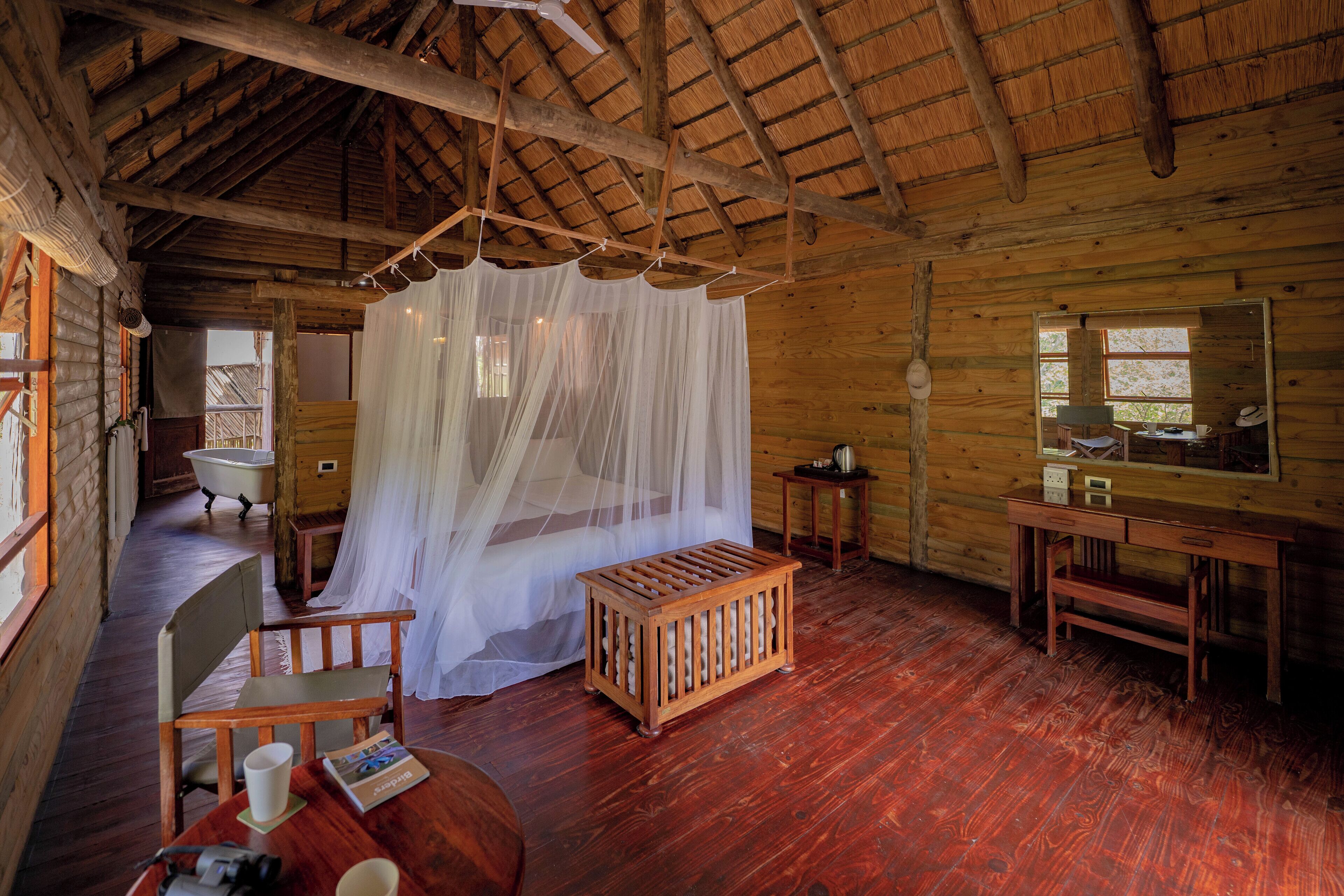 A rustic wooden cabin with a thatched roof, a wooden bench, a table with a magazine, a bed with a canopy, and a small wooden cabinet.